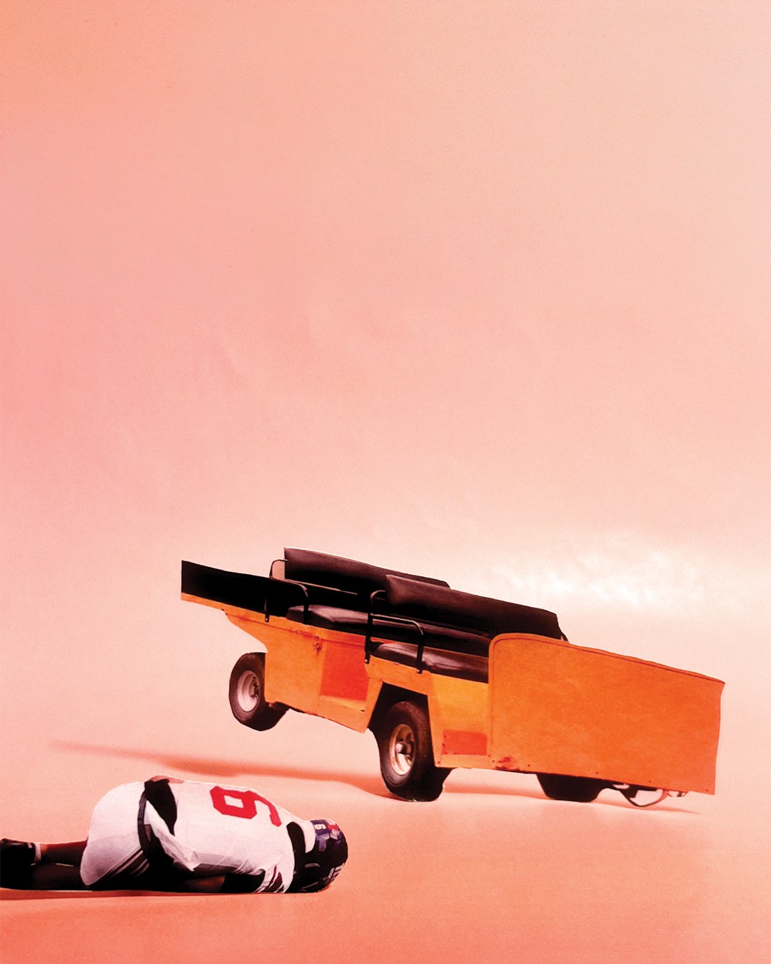 An empty orange injury cart tilted in the air against a peach background, with a player in a white number 6 jersey lying face down on the ground in front of it.