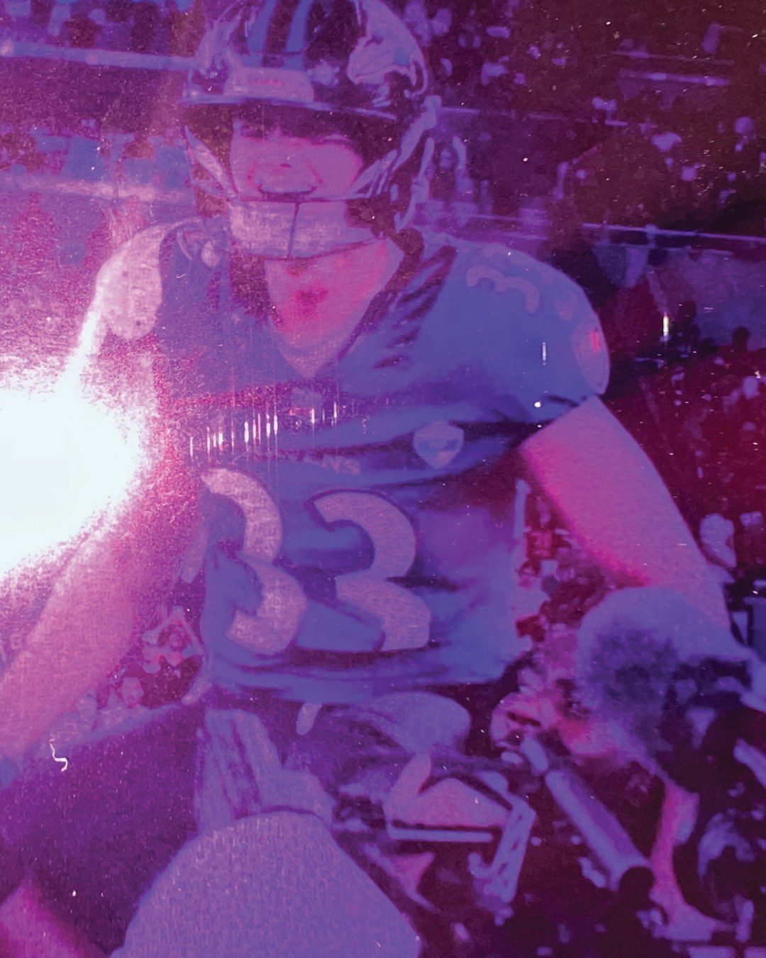 Ravens kicker number 33 on the sideline, lit by a burst of white light, in the same purple and magenta grain.